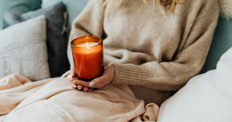 Une femme dans son le canapé, sous le plaid, tenant une bougie parfumée artisanale allumée. Guide complet bougies parfumées artisanales en cire végétale — N'importe Où